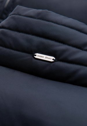 Niebieska kurtka puchowa z pikowanym wzorem, z metalową metką z napisem "OMBRE EXTIMO" oraz wzmocnionymi szwami.