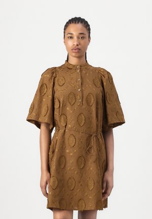 Femme aux cheveux tressés portant une robe marron avec des manches bouffantes, des motifs brodés, un col boutonné et une cordelette nouée à la taille.