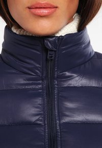 Granatowa pikowana kamizelka z wysokim kołnierzem, zapinana na zamek z przodu i z prążkowanym wnętrzem. Gładka faktura z przeszyciami na panelach.