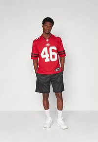 Raudonas futbolo marškinėliai su juodais akcentais ir baltu skaičiumi 46, derinami su juodais tinkliniais šortais ir baltais sportiniais bateliais.