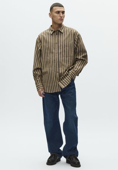 Młody mężczyzna w brązowej koszuli w paski z długim rękawem, luźnych niebieskich dżinsach i czarnych butach, stojący z jedną ręką w kieszeni.