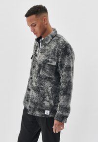 Jachetă din fleece cu model gri și negru, cu guler, butoane frontale, două buzunare la piept și o etichetă pe tiv. Material texturat și cald.