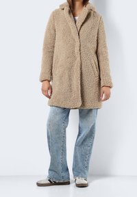 Cappotto teddy beige e peloso con colletto, maniche lunghe, tasche frontali e taglio fino al ginocchio, indossato sopra jeans di denim blu.