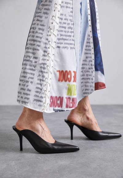 Mules de cuero negro con punta afilada y tacón de aguja, combinados con una prenda blanca con estampado que presenta detalles de cordones.