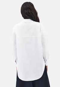 Frau mit langen dunklen Haaren, die ein locker sitzendes, langärmliges weißes Hemd und dunkle Hosen trägt, steht mit dem Rücken zu einem schlichten Hintergrund.