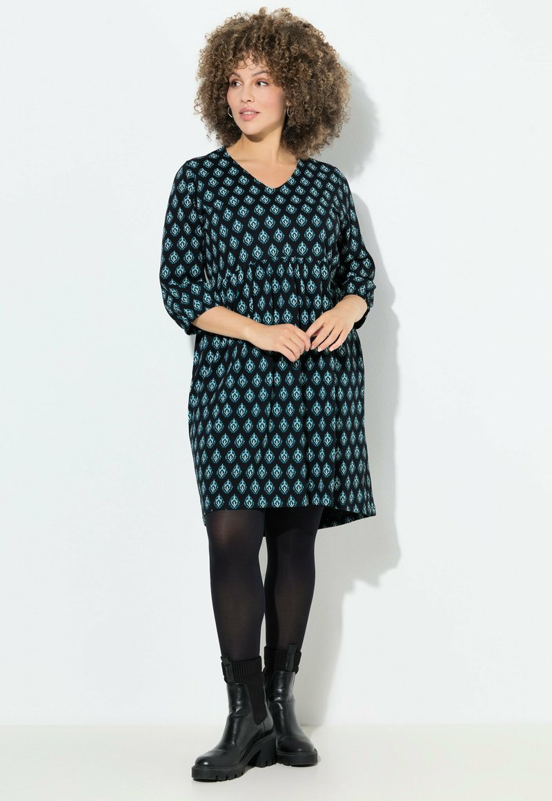 Musta mekko sinisellä geometrisella kuviolla, v-pääntie ja rypytetty vyötärö. Kolme neljäsosaa hihat. Mustat legginssit ja tukevat mustat saappaat viimeistelevät ilmeen.