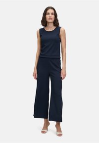 Marineblauwe jumpsuit met een mouwloze top en wijde pijpen, gemaakt van zachte stof. Heeft een relaxte pasvorm en een elastische tailleband, gecombineerd met lichte hakken.