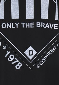 Schwarzes T-Shirt mit einem grauen grafischen Design mit vertikalen Streifen und fettem Text "NUR DIE MUTIGEN". Es enthält Markenakzente und ein Logo.