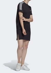 Personne portant une robe T-shirt noire d'Adidas avec des rayures blanches sur les manches et des baskets blanches, se tenant devant un fond uni.