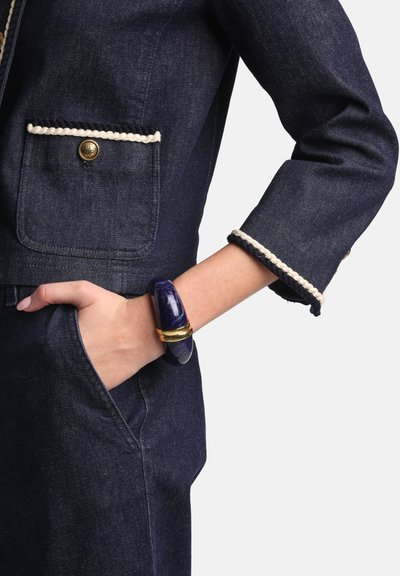 Denim jacket with gold buttons, rope trim on pockets, and an ornate bracelet featuring purple and gold accents worn on the wrist.