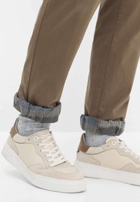 Sneaker in suede e pelle beige con suole bianche, abbinate a pantaloni marroni arrotolati per rivelare calzini grigi e un polsino a fantasia blu e grigio.