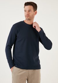 Tengerészkék hosszúujjú póló ribbed textúrával, kerek nyakkivágással és laza fazonnal. Világos színű nadrággal párosítva.