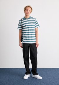 Striped teal and white cotton t-shirt with a round neckline, paired with black trousers and white sneakers, showcasing a casual look.