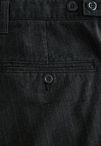 Dunkel gestreifter Stoff mit einer geknöpften Paspeltasche und zwei Knöpfen entlang des Taillenbunds eines maßgeschneiderten Kleidungsstücks.