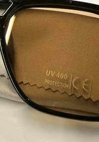 Slnečné okuliare s čiernym rámom ležia na hnedom mikrovláknovom utierke s nápisom „UV 400 Protection CE“, čo indikuje filtrovanie UV žiarenia a súlad s európskymi bezpečnostnými normami.