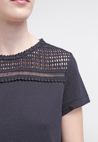 Tmavě šedé tričko s detailním krajkovým jhem s geometrickými vzory a vlnitým lemem, vyrobené z měkkého materiálu s krátkými rukávy.