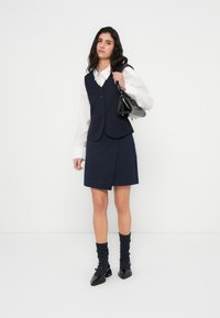 Μπλε γιλέκο πάνω από λευκό πουκάμισο, συνδυασμένο με φούστα σε ίδιο χρώμα. Μαύρη τσάντα και παπούτσια με μαύρες υφασμάτινες κάλτσες ολοκληρώνουν το σύνολο.