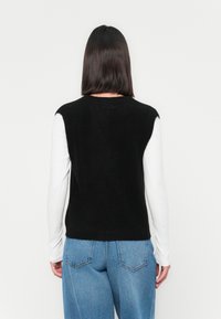 Persoon met schouderlang donker haar, gekleed in een zwarte mouwloze trui over een wit langemouwshirt en blauwe spijkerbroek, met de rug naar de camera gekeerd.