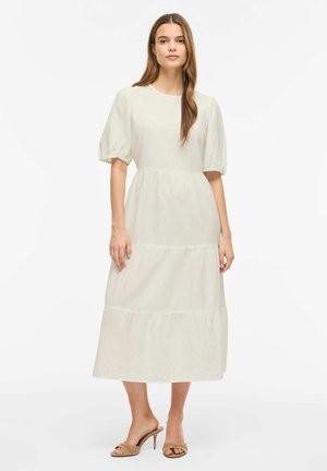 Femme portant une robe midi blanche avec des manches bouffantes et une jupe à volants, associée à des sandales à talons beiges à bout ouvert, debout devant un fond blanc.