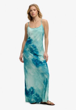 Kobieta w niebieskiej, długa do ziemi sukience tie-dye bez rękawów z cienkimi ramiączkami i brązowymi, płaskimi sandałami, stojąca na tle jednolitego tła.