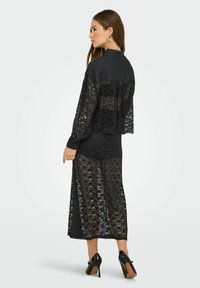 Robe en dentelle noire avec des manches longues et un col haut, présentant un design superposé, une texture semi-transparent et une silhouette fluide.