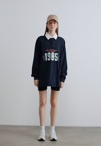 Μπλε σκούρο πόλο με μακριά μανίκια και λευκό γιακά, με σχέδιο γραφικού του 1985. Φορεμένο με μαύρο σορτς και λευκά αθλητικά παπούτσια, τοποθετημένο σε απλό φόντο.