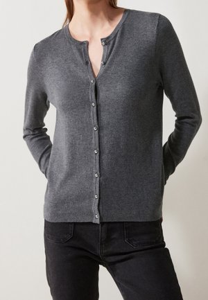 Vrouw draagt een nauwsluitend donkergrijs gebreid vest met knopen, lange mouwen en zwarte broek, tegen een neutrale achtergrond.