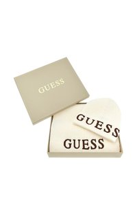 Conjunto de gorro y bufanda de punto blanco en una caja beige. Ambas piezas cuentan con el logo "GUESS" en marrón. Textura suave, diseño acanalado y forma redondeada del gorro.