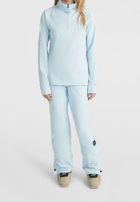 Kobieta ubrana w jasnoniebieską bluzę z długim rękawem i zamkiem błyskawicznym oraz pasujące spodnie narciarskie nad beżowymi butami narciarskimi, stojąca na białym tle.