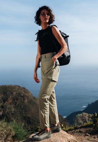 Pantaloni khaki leggeri, top senza maniche nero, scarpe grigie con dettagli arancioni e zaino nero. Ambientazione all'aperto con vista sull'oceano.