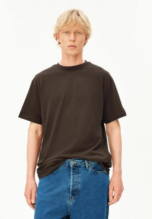 Blonde man met kort haar die een effen donkerbruin T-shirt en blauwe spijkerbroek draagt, staand tegen een witte achtergrond.