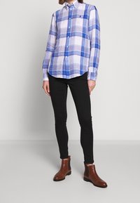 Camicia a quadri blu e bianca con bottoni abbinata a jeans skinny neri e stivaletti alla caviglia marroni. La camicia presenta un colletto e un dettaglio con logo.