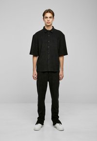 Camisa negra de mangas cortas con botones combinada con pantalones negros con cordón. La tela parece suave y ligeramente texturada. El modelo está de pie frente a un fondo simple.