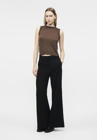 Γυναίκα που στέκεται με το ένα χέρι στην τσέπη, φοράει αμάνικο καφέ τοπ και παντελόνες με φαρδιά μπατζάκια σε μαύρο χρώμα, με μαύρα παπούτσια, σε απλό φόντο.