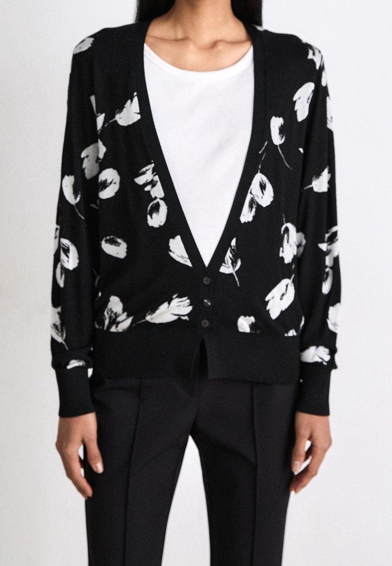Personne portant un cardigan noir à imprimé floral blanc sur un t-shirt blanc et un pantalon noir, debout devant un fond clair uni.