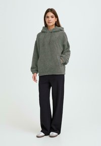 Grüner, flauschiger Kapuzenpulli mit Kordelzügen und Seitentaschen, kombiniert mit schwarzen, weit geschnittenen Hosen und grauen Hausschuhen. Einfaches, gemütliches Design.