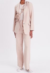 Veste de costume beige ajustée et pantalon à jambes larges, haut texturé, associé à un sac bandoulière beige et des baskets beiges. Accents en métal argenté.