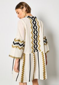 Robe crème avec des manches cloche, un motif zigzag cousu en or, noir et blanc, et un design de jupe à volants.