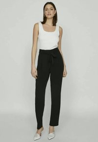 Sieviete stāv, ģērbusies baltā bezpiedurkņu topā, augstas jostas melnās biksēs ar sasaistītu jostu un baltos uzgurnus apavus ar rotājumiem.