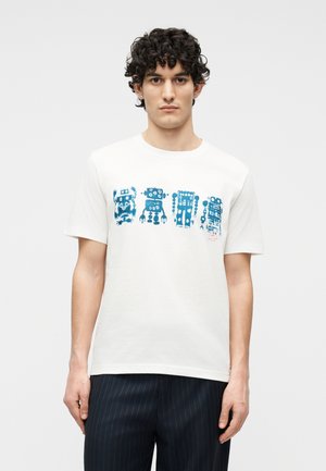 Giovane uomo con capelli ricci e scuri che indossa una maglietta bianca con una stampa di un robot blu sul petto e pantaloni scuri a righe sottili.