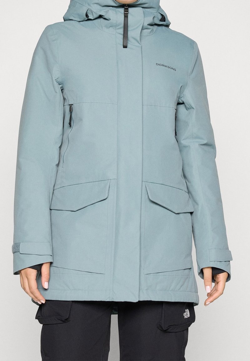 Personne portant une veste d'hiver à capuche Didriksons bleu clair avec poches avant et un pantalon cargo noir.