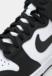 Černobílé tenisky s hladkým koženým svrškem, černými tkaničkami, perforovanou špičkou a výrazným černým swooshem na boku.