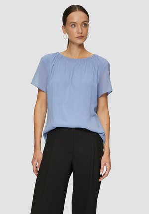 Hellblaues Kurzarmbluse mit gerafftem Ausschnitt, luftiger Silhouette und weichem Stoff, kombiniert mit schwarzen hochgeschnittenen Hosen.