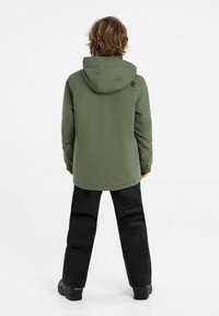 Veste rembourrée verte avec capuche, dotée de poignets élastiques, de poches latérales et d'un logo dans le dos, portée avec un pantalon noir et des bottes.