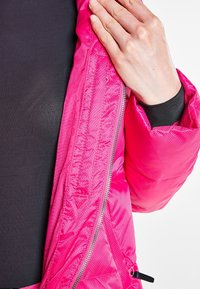 Doudoune fuchsia avec une finition brillante, dotée d'une fermeture éclair et d'un tissu texturé. La manche possède une bande élastique pour un ajustement confortable.