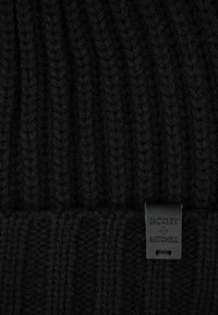 Schwarze Strickmütze mit vertikal geribbelter Textur, ausgestattet mit einem Lederetikett, auf dem "BICKLEY + MITCHELL" geprägt und am Rand angenäht ist.