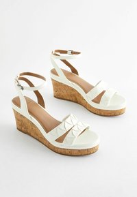 Sandales blanches à talon compensé en liège, avec deux larges brides à bords festonnés et une bride à la cheville avec une boucle. Fini en cuir lisse.