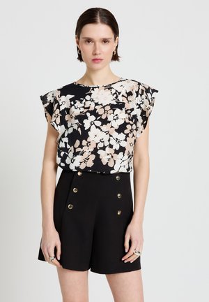 Frau trägt eine schwarze Blumenkurzarmbluse und hoch taillierte schwarze Shorts mit goldenen Knöpfen vor einfarbigem Hintergrund.