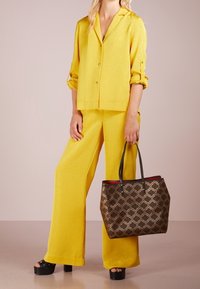 Żółta bluzka o fakturze i szerokie spodnie z szerokimi nogawkami, zestawione z ciemną torbą typu tote z wzorem, z czarnymi uchwytami i czerwonym wnętrzem. Sandały na platformie.