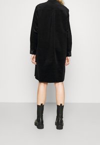 Czarna sukienka koszulowa z kordurowego materiału, z długimi rękawami i zapięciem na guziki, o prostym kroju i detalem szwu z tyłu. Połączona z czarnymi botkami do kolan.